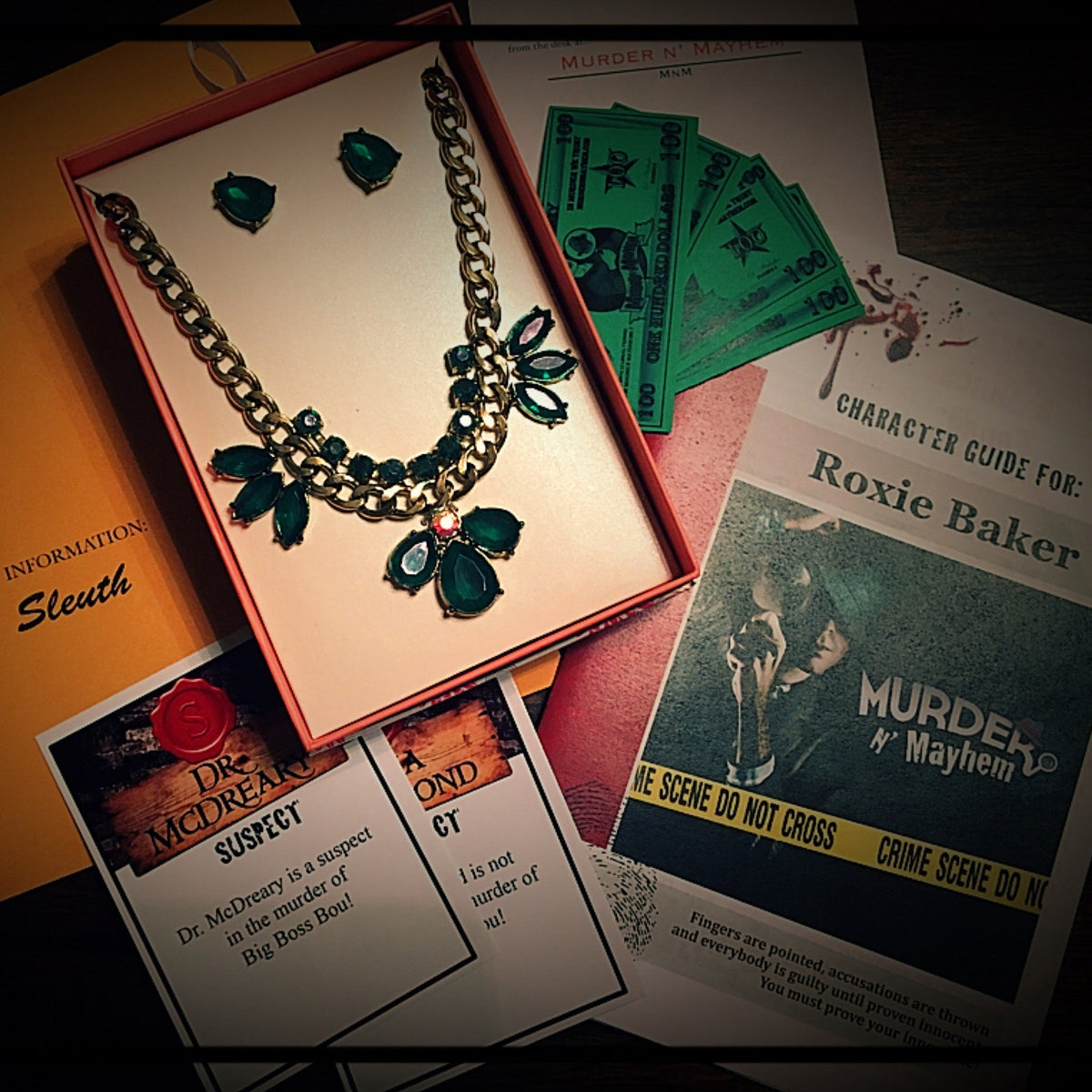 A gothic-style jewelry set featuring a silver-tone chain necklace and matching earrings with dark green marquise-cut stones and a small red accent, displayed in an open red box. Surrounding props include fake money, character guide cards, and a magazine on a dark wooden surface.